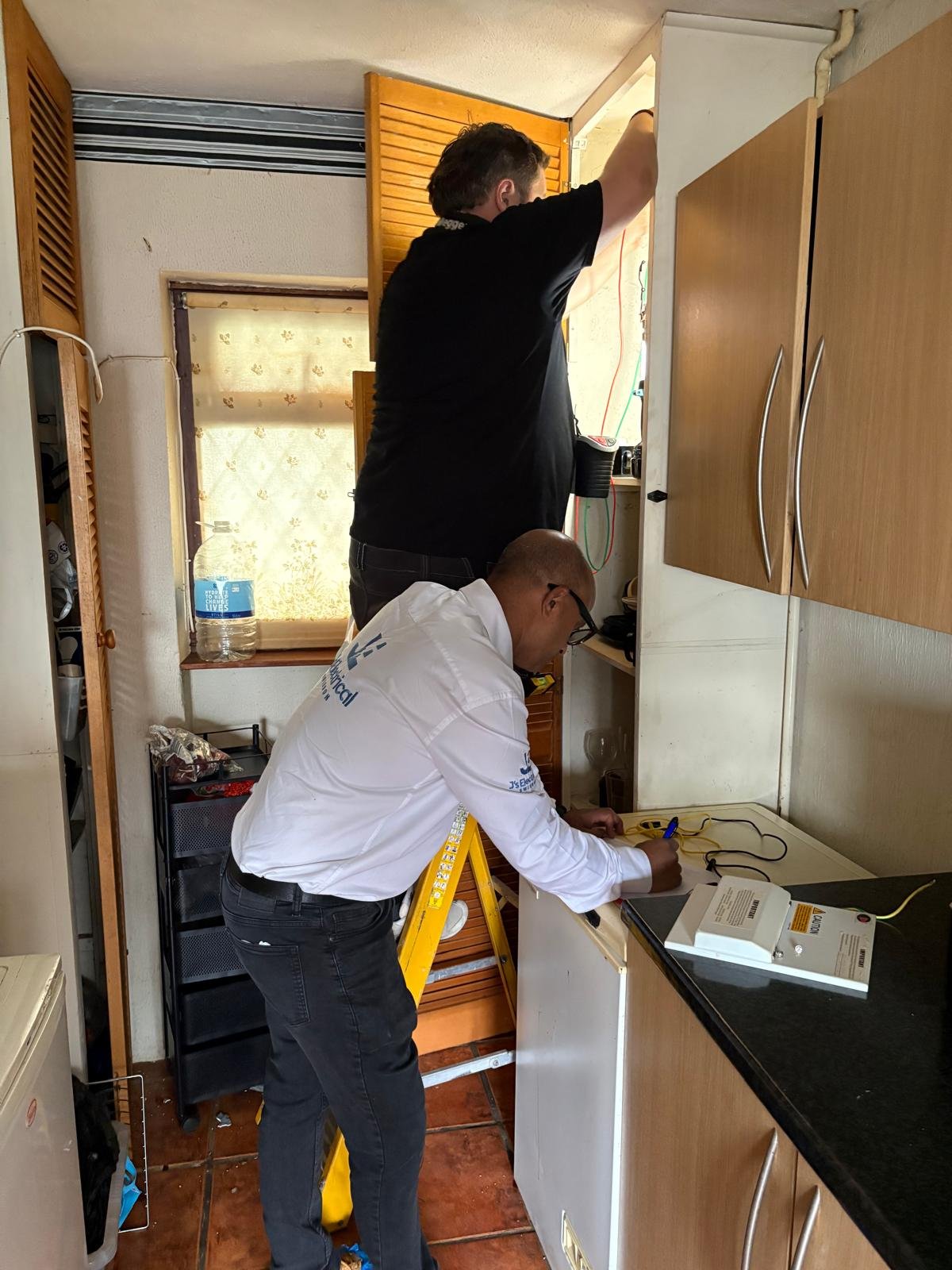 Junab Ali writing up test results while a colleague works inside a cupboard during an electrical inspection